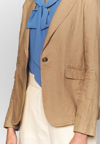 Blazer testardo marrone chiaro, con texture e un solo bottone, abbinato a una blusa blu con dettaglio a fiocco. L'outfit comprende una gonna color crema.