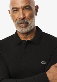 Schwarzes Langarm-Polohemd aus strukturiertem Stoff, mit drei Knöpfen und einem kleinen gestickten Krokodil-Logo auf der Brust.