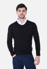 Giovane uomo con capelli e barba scuri, che indossa un maglione nero a V sopra una camicia bianca e jeans blu, in piedi con una mano in tasca.