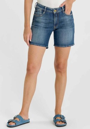 Vrouw met blauwe denimshorts en blauwe instapsandalen staat tegen een effen achtergrond.