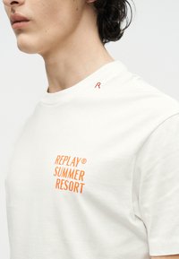 Νεαρό άτομο που φοράει λευκό μπλουζάκι με πορτοκαλί κείμενο "REPLAY SUMMER RESORT" στο στήθος και μικρό κόκκινο "R" κοντά στον ώμο.