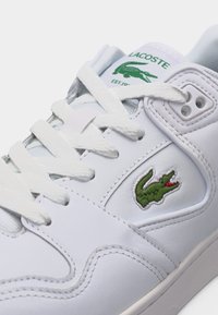 Λευκό αθλητικό παπούτσι Lacoste με πράσινο εικονίδιο κροκοδείλου στο πλάι και τη γλώσσα, με λευκά κορδόνια και λεπτομερείς ραφές.