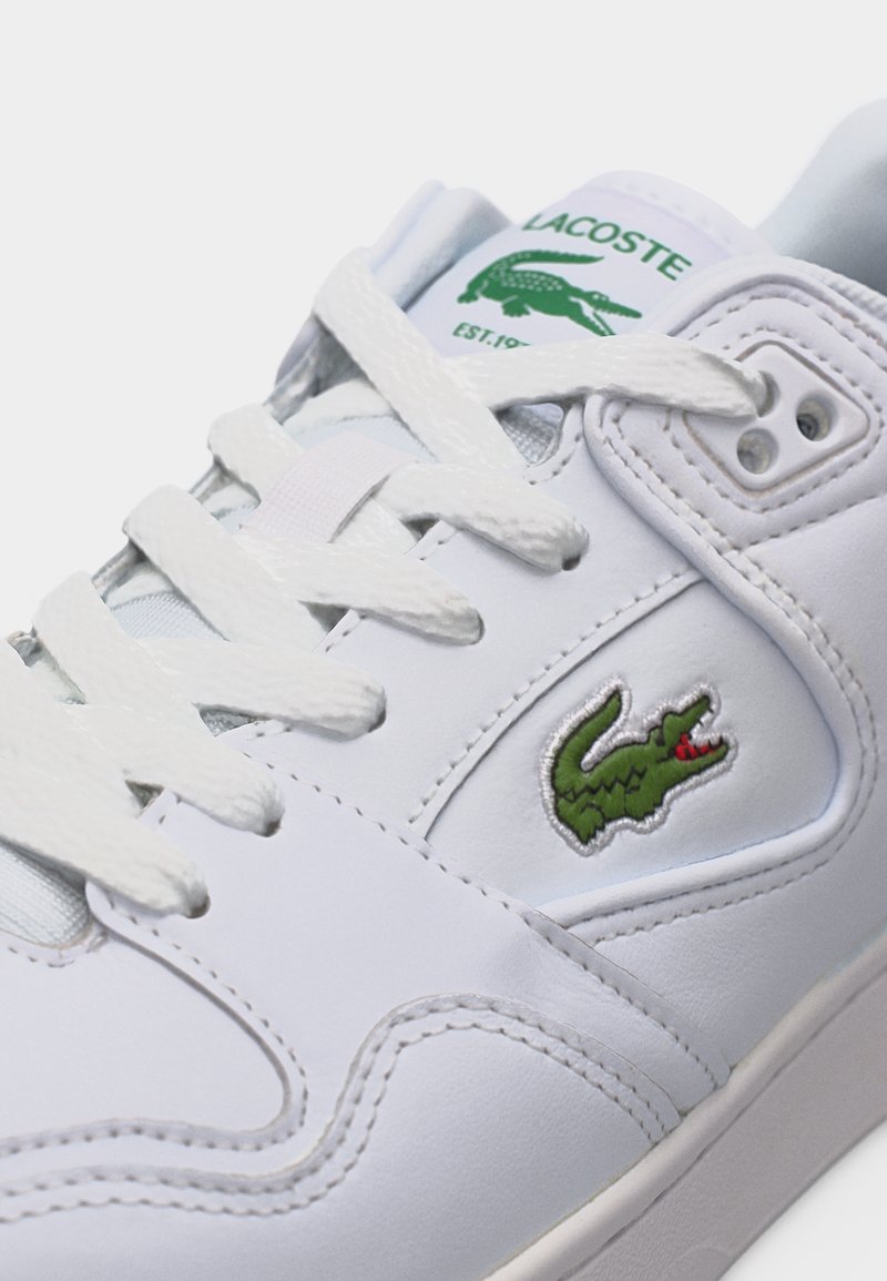 Witte Lacoste-sneaker met groen krokodillenlogo op de zijkant en tong, voorzien van witte veters en gedetailleerde stiksels.