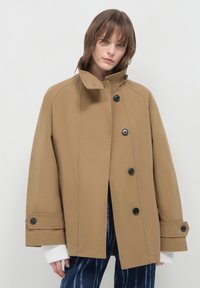 Frau trägt einen übergroßen beigen Knopfmantel mit hohem Kragen, die Ärmel eines weißen Pullovers sind sichtbar, und sie trägt eine Hose mit blauem Muster vor einem einfarbigen Hintergrund.