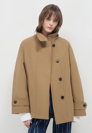 Donna che indossa un cappotto beige oversize con bottoni e colletto alto, maniche di maglione bianco visibili, e pantaloni blu a fantasia su sfondo neutro.