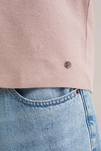 Gaiši rozā teksturēts tops ar apaļu apakšmalu, kas rotāts ar mazu logotipa piespraudi. Kombinācijā ar klasiskām zilām džinsu biksēm, kurām redzami kabatu detaļas.