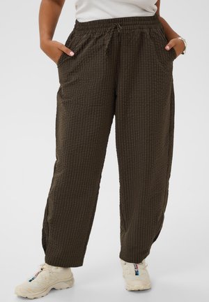 Personne portant un pantalon ample marron foncé à carreaux avec taille élastique et baskets blanches, mains dans les poches sur un fond uni.