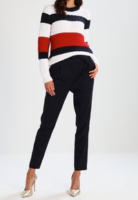 Stickad tröja med vita, marinblå och röda horisontella ränder, texturerat mönster; matchad med marinblå skräddarsydda byxor och guldklackar.