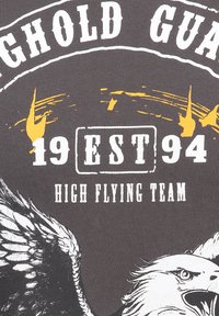 Camiseta gris con un estampado gráfico de un águila, texto blanco "EQUIPO DE ALTAS VUELOS" y acentos amarillos desgastados. Tamaño y año "EST 94" exhibidos.