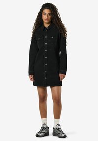 Mladá žena s dlouhými kudrnatými vlasy, oblečená v černých džínových šatech na knoflíky se stříbrnými hroty a stříbrných teniskách s bílými ponožkami.