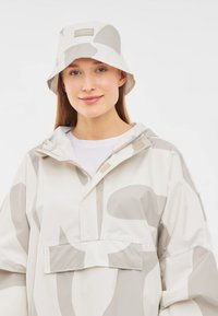 Ľahká, béžová pršiplášť s abstraktnými šedými vzormi, prednou vreckom a kapucňou, doplnená zodpovedajúcim béžovým klobúkom.