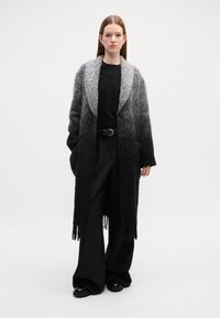 Cappotto lungo in lana grigio sfumato a nero con una finitura testurizzata, ampi revers e orlo frangiato; abbinato a pantaloni e scarpe neri.
