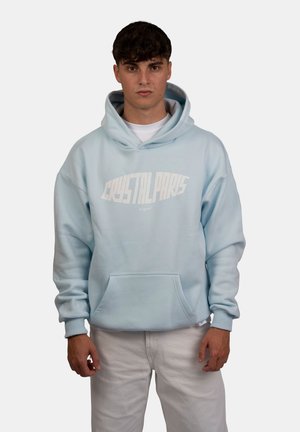 Felpa leggera di colore azzurro con una grande scritta grafica bianca "CRYSTAL PARIS" sul davanti, con tasca a marsupio e polsini a coste, indossata sopra pantaloni bianchi.