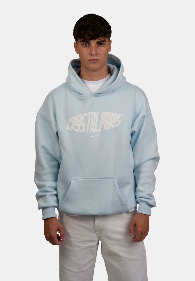 Felpa leggera di colore azzurro con una grande scritta grafica bianca "CRYSTAL PARIS" sul davanti, con tasca a marsupio e polsini a coste, indossata sopra pantaloni bianchi.
