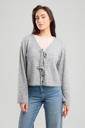 Cardigan corto grigio chiaro in morbido maglione, con scollatura a V, chiusura con lacci sul davanti e maniche lunghe. Indossato sopra una maglietta in pizzo.