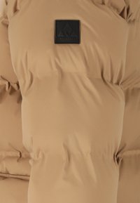 Beige pufferjakke med quiltet design. Sort stoflogo på øverste venstre side. Materialet ser glat og isoleret ud.