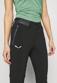 Černé turistické kalhoty s texturovaným černým pasem, vybavené boční kapsou na zip a bílým logem na stehně.