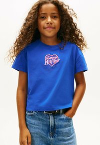 Camiseta de algodón azul con mangas cortas, con un logo bordado en forma de corazón en rosa y blanco, combinada con un denim azul claro.