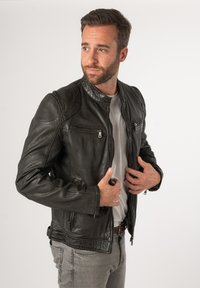 Schwarze Lederjacke mit Reißverschlusstaschen, strukturierten Schulterdetails und slim fit Design. Getragen über einem hellgrauen Hemd.