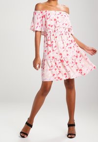 Vestido off the shoulder en rosa claro con estampado de hojas rojas, mangas con volantes, cintura ajustada y dobladillo acampanado, combinado con tacones negros con tiras.