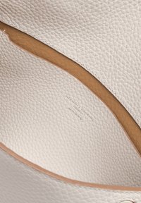 Interno di una borsa a tracolla bianca in pelle, caratterizzato da una superficie testurizzata, foderatura liscia color mogano e un delicato marchio impresso sul fondo.