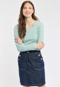 Femme aux longs cheveux bruns portant un haut à manches longues bleu clair et une jupe en denim foncé avec des détails de boutons, debout les bras croisés.