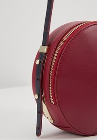Borsa rotonda in pelle rossa con zip dorata, manico blu e dettagli dorati. Texture liscia con design minimalista.