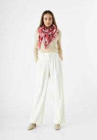 Femme stand in les poches des pantalons larges blancs, pull beige, écharpe à motifs roses et bottines beiges sur fond blanc.