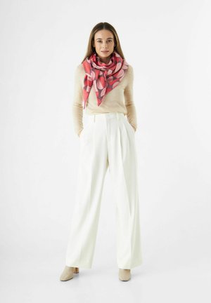 Femme stand in les poches des pantalons larges blancs, pull beige, écharpe à motifs roses et bottines beiges sur fond blanc.