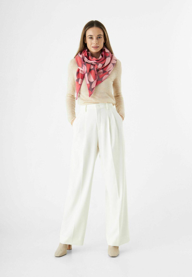 Femme stand in les poches des pantalons larges blancs, pull beige, écharpe à motifs roses et bottines beiges sur fond blanc.