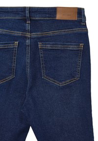 Jeans in denim blu scuro con una consistenza liscia, caratterizzati da cuciture gialle e una toppa in pelle marrone alla vita. Presentano due tasche posteriori.