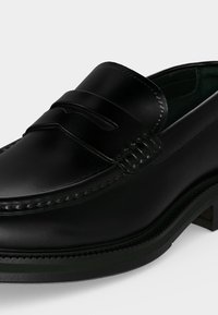Chaussure penny loafer en cuir noir avec des détails cousus et une finition lisse sur un fond blanc.
