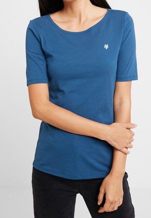 Vrouw draagt een blauw T-shirt met korte mouwen en een klein wit logo op de borst, gecombineerd met een zwarte broek, staat met gekruiste armen.