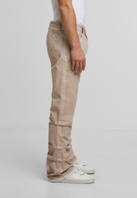 Mies, joka seisoo vaaleissa housuissa ja valkoisissa lenkkareissa, yksinkertaista vaaleanharmaata taustaa vasten, sivuprofiili, joka näyttää rennon istuvuuden ja poimuttuneen kankaan.