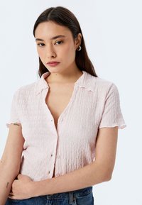 Blusa rosa chiaro a maniche corte con tessuto testurizzato, dettagli con bottoni e colletto a punta. Indossata con jeans a vita alta blu.