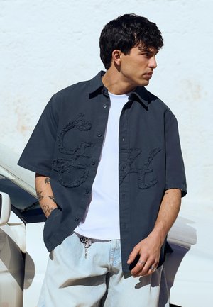 Jonge man met kort donker haar draagt een marineblauw overhemd met knopen over een wit T-shirt, lichte jeans en zilveren ringen, staand bij een witte auto.