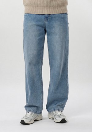 Person trägt hellblaue Jeans mit geradem Bein und weiße New Balance-Sneaker, steht vor einem schlichten weißen Hintergrund.