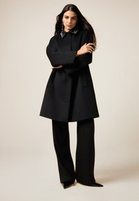 Abrigo de lana negro con cuello de solapa que presenta elementos decorativos, combinado con pantalones negros rectos y zapatos de punta afilada.