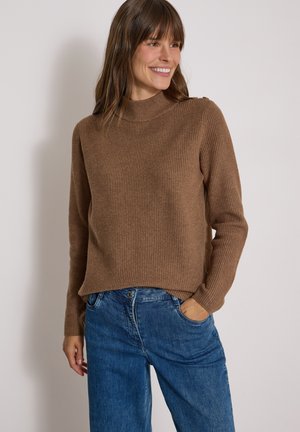 Femme souriante portant un pull côtelé marron avec des détails de boutons sur l'épaule et un jean bleu, debout avec une main dans la poche.