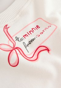 Tessuto bianco con testo stampato in rosso e nero: "a: Minnie da: Babbo Natale", con un design di un nastro rosa e un bordo rettangolare.