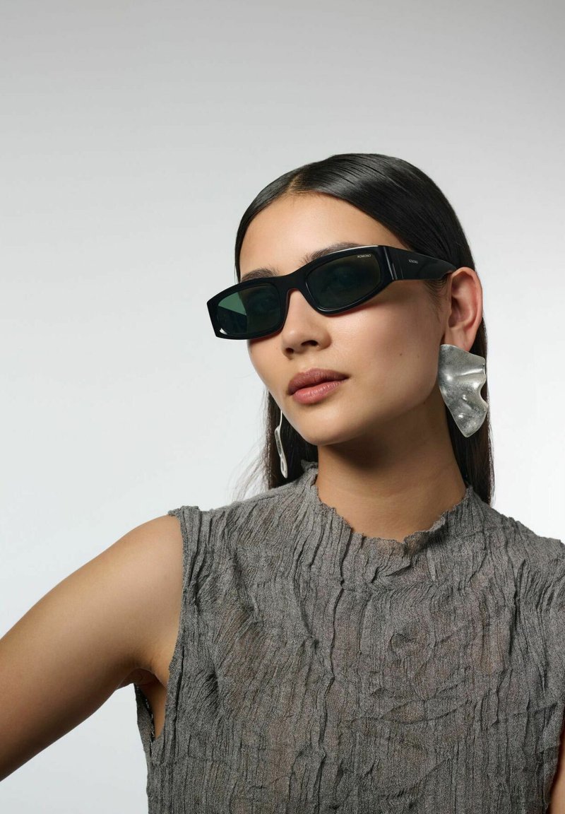 Femme aux longs cheveux noirs portant des lunettes de soleil noires angulaires, de grandes boucles d'oreilles argentées abstraites et un haut gris texturé sans manches, sur un fond uni.