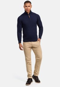 Marineblauer Kabelstrickpullover mit einem Halb-Reißverschluss-Kragen, kombiniert mit beigen Hosen. Das Outfit besticht durch einen strukturierten Stoff und dunkelbraune Schuhe.