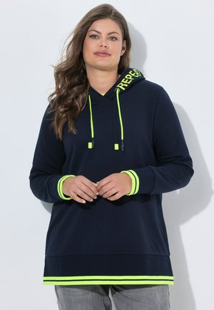 Kvinde iført en marineblå hoodie med neon-gule snore, manchet- og kantstriber og teksten "REPEAT" på hætten, der står foran en hvid baggrund.
