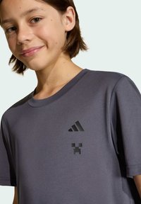 Tmavošedé tričko s krátkymi rukávmi a okrúhlym výstrihom, s čiernym logom Adidas a pixelovou grafikou na ľavej strane hrude.