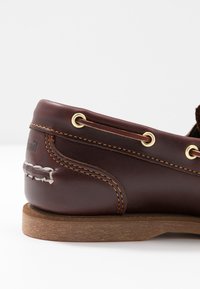 Braune Lederslipper mit goldenen Ösen, kontrastierendem gelbem Stich und einer dunkelbraunen flachen Sohle. Verfügt über dekorative Schnürdetails.