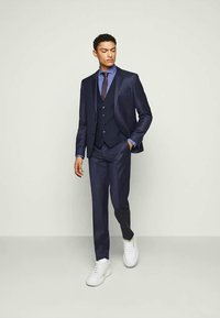 Hombre con traje de tres piezas azul marino, camisa azul, corbata marrón, caminando con una mano en el bolsillo, llevando zapatillas blancas sobre un fondo liso.