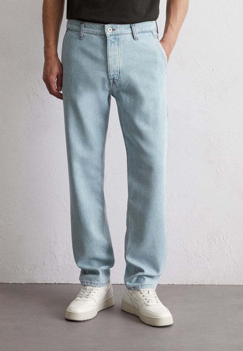 Jasnoniebieskie dżinsy w prostym kroju, z pięcioma kieszeniami i kontrastowym przeszyciem. W połączeniu z białymi trampkami na neutralnym tle.