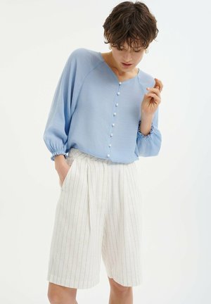 Light blue button-up blouse with puffy sleeves and accent buttons, paired with cream pinstriped shorts. Soft fabric texture; relaxed fit.