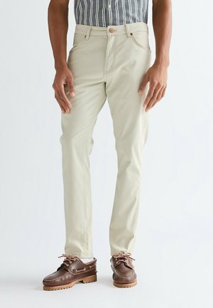 Mann trägt beige Hose, gestreiftes Hemd eingesteckt und braune Leder-Schnürschuhe, steht vor einfachem Hintergrund.