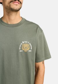Olijfgroene katoenen t-shirt met een grafische afbeelding van een leeuwenhoofd en tekst "RVCA EST. MMI" in crème en goud, met een klassiek rond decolleté ontwerp.